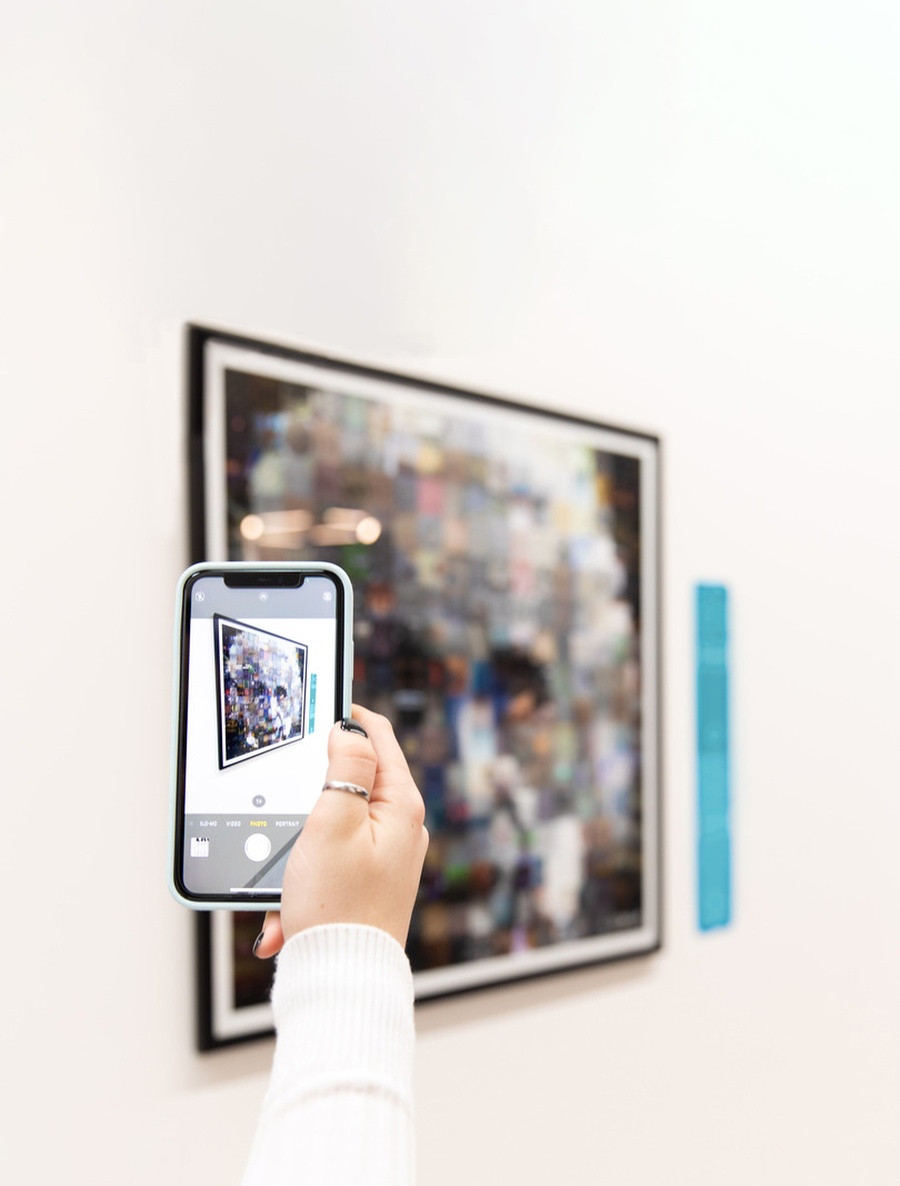 A hand is holding a phone, capturing the Master of Numbers exhibit on the wall at Museum of Illusions Manchester. The exhibit shows Albert Einstein's face composed of numerous small images and is an artwork by Gianni Sarcone.