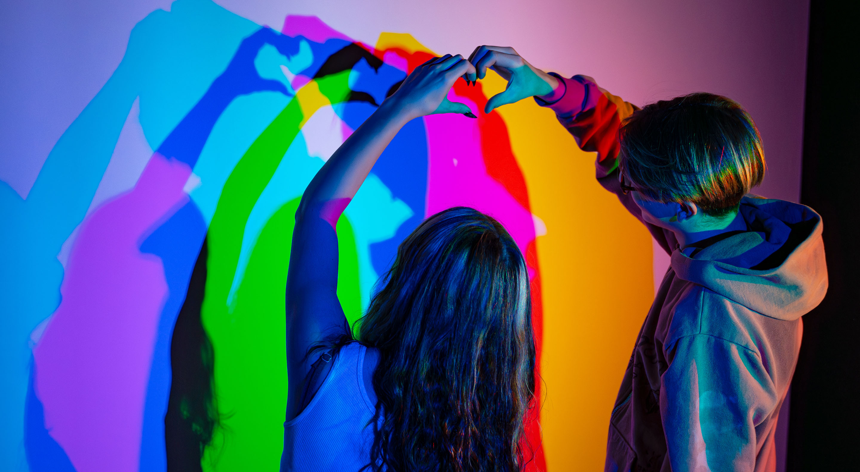 Young couple in the Colour Room creates a heart with their hands, while casting colorful shadows on the wall.